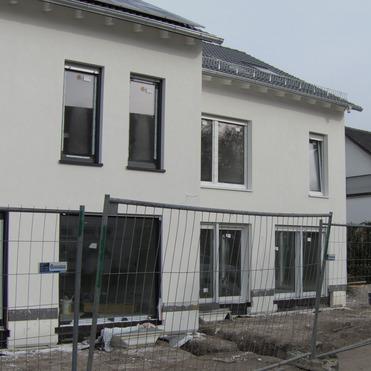 Neubau eines weißen Hauses mit Baustellenzaun und unbefestigtem Boden an einer Straße.