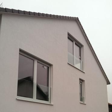 Fassade eines modernen Hauses mit großen Fenstern und Balkon, unvollendetes Grundstück im Vordergrund.