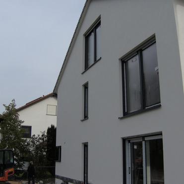 Modernes, unvollendetes Haus mit großen Fenstern, daneben ein Bagger und Gartenbereich in einer Wohngegend.