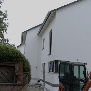 Blick auf ein Wohnhaus mit weißen Wänden, einem Bagger und einem bepflanzten Holzzaun im Vordergrund.
