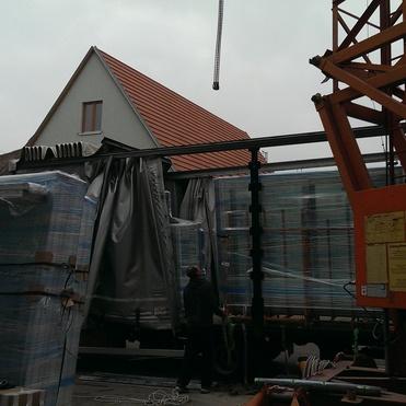 Lkw-Ladefläche mit verpackten Materialien und einem Kranarm, im Hintergrund ein Wohnhaus und bewölkter Himmel.
