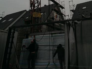 Baukran hebt Material über einen Lastwagen, während Arbeiter die Ladung sichern, vor grauem Himmel und Baustellenkulisse.