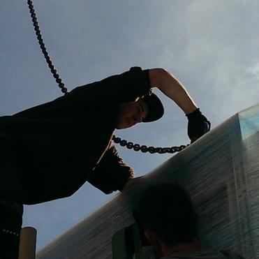 Arbeiter hebt eine große, verpackte Ladung an einem Kran mit einer Kettenmechanik an.