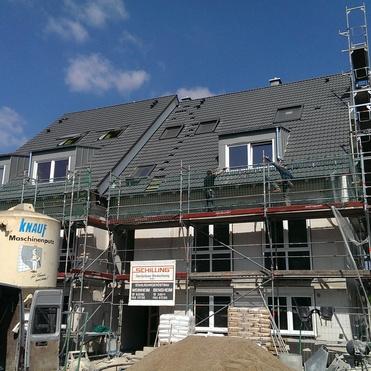 Bau eines Mehrfamilienhauses mit Gerüst, noch nicht vollständig gedecktem Dach und Baustellenfahrzeugen im Vordergrund.