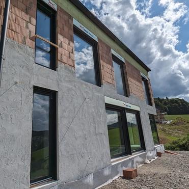Fertighaus im Bau mit großen Fenstern und unvollständiger Fassade, umgeben von grünen Wiesen und wolkigem Himmel.