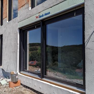 Baufortschritt eines Hauses mit großen Fenstern und grünem Dach, umgeben von einer unbebauten Landschaft.