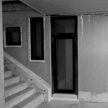 Eingangstür aus Glas in einer Baustelle, umgeben von unvollendeten Wänden und einer Treppe.