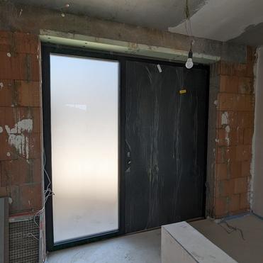 Blick auf eine Baustelle mit einer schwarzen Glastür und mattem Glas neben unverputzten Wänden und einem Betonboden.