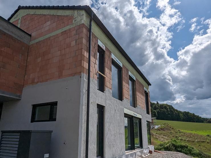 Baustelle eines zweigeschossigen Hauses mit unvollständig verputzter Fassade, umgeben von einer grünen Wiese und Wolken.