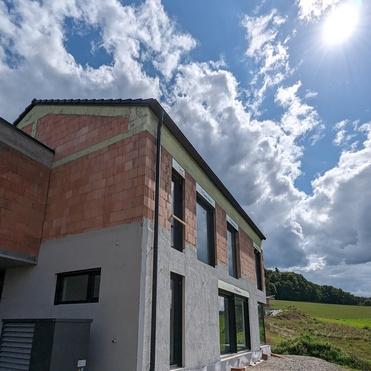 Baustelle eines zweigeschossigen Hauses mit unvollständig verputzter Fassade, umgeben von einer grünen Wiese und Wolken.