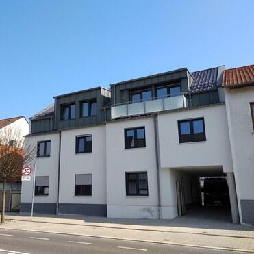 Moderne Wohnanlage mit flachdachigem Obergeschoss und Balkon, an einer Stadtstraße mit Nachbarhäusern.