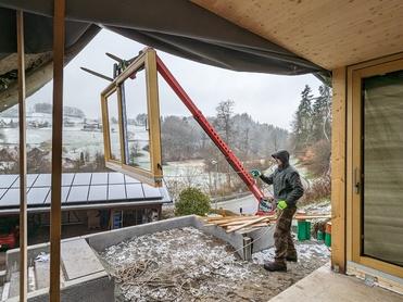 Zwei Bauarbeiter montieren ein großes Fenster mit einem Kran in ein neues Gebäude bei winterlichem Wetter.