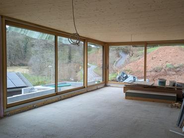 Innenraum eines unvollendeten Hauses mit großen Fenstern und Blick auf eine grüne Landschaft. Baustellenmaterial liegt verstreut.