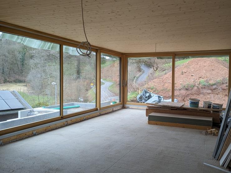 Innenraum eines unvollendeten Hauses mit großen Fenstern und Blick auf eine grüne Landschaft. Baustellenmaterial liegt verstreut.