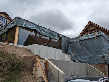 Bauprojekt mit grau abgedecktem Anbau, Sicht auf Treppe und Wohnhäuser im Hintergrund unter bewölktem Himmel.