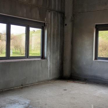 Blick in einen ungeklärten Raum mit zwei Fenstern, grauen Wänden und einem nackten Betonboden.