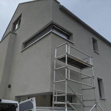 Neues Wohnhaus mit grau verputzter Fassade und Gerüst an der Ecke für Malerarbeiten. Wolkenverhangener Himmel im Hintergrund.