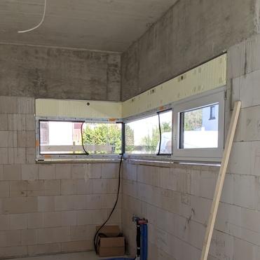 Leere, unfertige Innenraumansicht mit zwei Fenstern, blauen Rohren und einem Holzstab an einer Wand.
