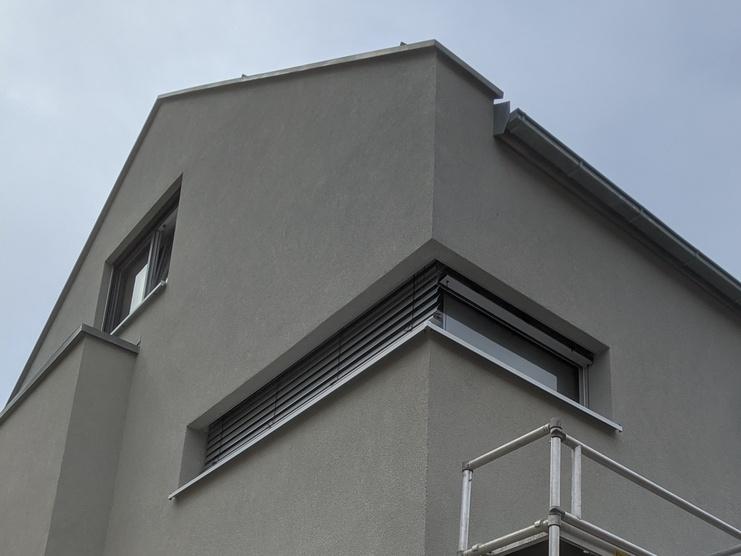 Neues Wohnhaus mit grau verputzter Fassade und Gerüst an der Ecke für Malerarbeiten. Wolkenverhangener Himmel im Hintergrund.