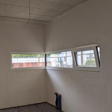 Leeres Zimmer mit grauem Boden, weißen Wänden und zwei schmalen Fenstern. Kabel hängen von der Decke.