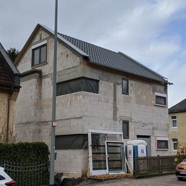 Blick auf ein im Bau befindliches Haus mit grauem Dach und unvollständiger Fassade an einer Straße mit parkenden Autos.
