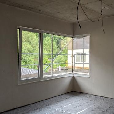 Unfertiger Raum mit grauem Teppichboden und großen Fenstern, die auf einen grünen Außenbereich blicken.