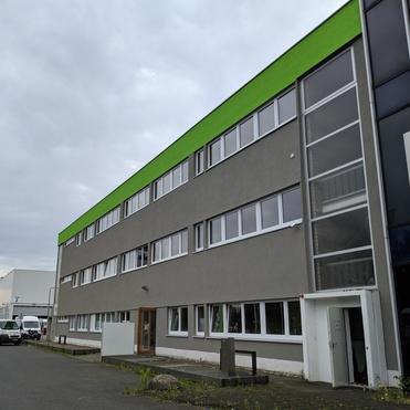 Moderner grauer Bürogebäudekomplex mit grünem Dach und großen Fenstern an einer unbefahrenen Straße.