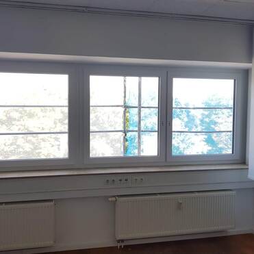 Blick auf drei Fenster mit grauen Fensterrahmen und Heizkörpern darunter, dahinter ist ein heller Außenbereich zu sehen.