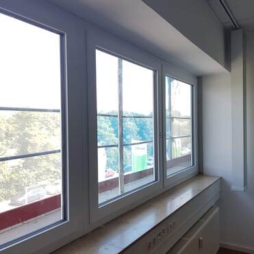 Vier große Fenster in einem hellen Raum mit Blick auf Bäume und einen Parkplatz im Freien.