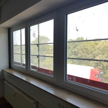 Blick aus einem Bürofenster auf eine grüne Baumlandschaft und einen Lkw auf einem Parkplatz. Fensterscheiben leicht verschmutzt.