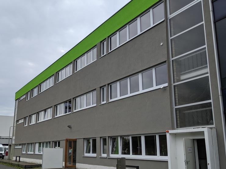 Moderner grauer Bürogebäudekomplex mit grünem Dach und großen Fenstern an einer unbefahrenen Straße.