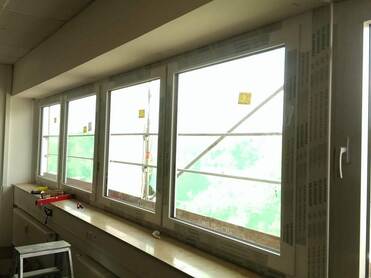 Blick auf mehrere Fenster mit Holzrahmen in einem Raum, Baugerüst und grüne Abdeckung im Hintergrund.