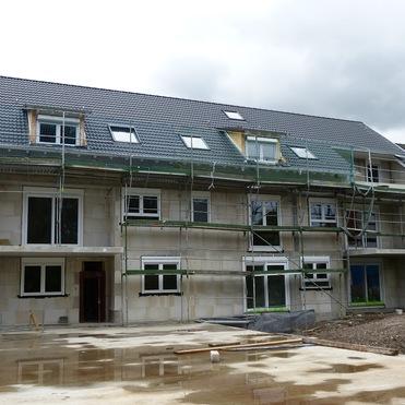 Baustelle eines mehrstöckigen Gebäudes mit Gerüst und verkleideten Fenstern, umgeben von unbefestigtem Boden.