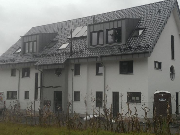 Neubau eines zweigeschossigen Wohnhauses mit grauem Dach und weißen Wänden, umgeben von Baufahrzeugen und ungestutztem Gras.