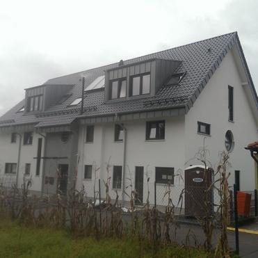 Moderne, zweigeschossige Wohngebäude mit Satteldach und großen Fenstern, umgeben von blühendem Gras und Nachbarhäusern.