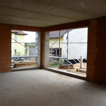 Blick durch ungefertigte Fenster in einen Rohbau, umgeben von Baustellenmaterial und benachbarten H&auml;usern.