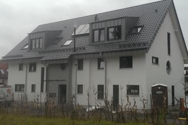 Neubau eines zweigeschossigen Wohnhauses mit grauem Dach und weißen Wänden, umgeben von Baufahrzeugen und ungestutztem Gras.