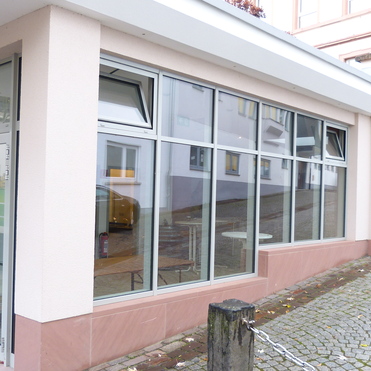Moderne, verglaste Au&szlig;enseite eines Geb&auml;udes mit eckigen Fenstern und einer gepflasterten Stra&szlig;e im Vordergrund.