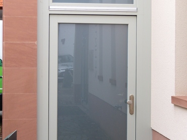 Glasierte Eingangst&uuml;r mit grauem Rahmen, umgeben von hellen W&auml;nden und einem gepflasterten Gehweg.