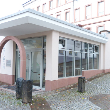 Fassade eines modernen Geb&auml;udes mit Glasfront und geschwungener Eingangst&uuml;r, in einer gepflasterten Stra&szlig;e.